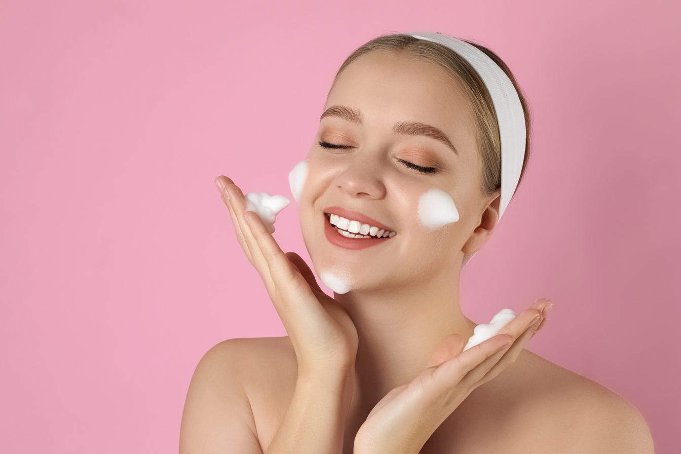 Young Woman Washing Face with Cleansing Foam on Pink Background.